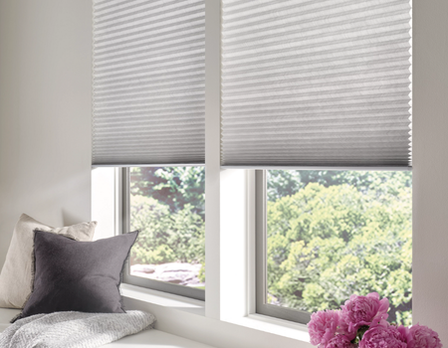 Window with white blinds and a view of greenery outside, with pillows and flowers on a windowsill.